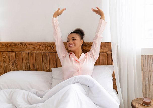 Jó reggelt, világ. Jól kipihent afro-amerikai nő ül és nyújtózkodik kényelmes ágyban, felemeli a kezét és mosolyog - Fotó, kép