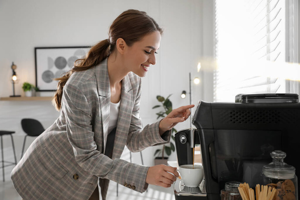 Νεαρή γυναίκα ετοιμάζει φρέσκο αρωματικό καφέ με σύγχρονη μηχανή στο γραφείο - Φωτογραφία, εικόνα