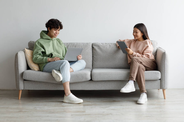 Schöne junge asiatische Kerl und seine Freundin mit modernen Gadgets, während sie auf der Couch zu Hause sitzen, Kopierraum - Foto, Bild