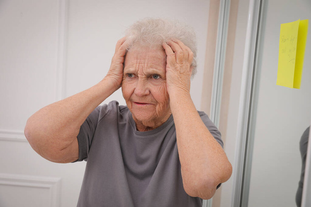Forgetful senior woman near mirror with reminder note indoors. Age-related memory impairment - Foto, Imagem