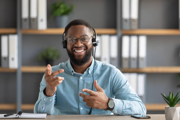 Χαμογελώντας φιλικό νεαρός αφροαμερικανός γενειοφόρος τύπος σε γυαλιά και ακουστικά χειρονομίες και κοιτάζει κάμερα - Φωτογραφία, εικόνα
