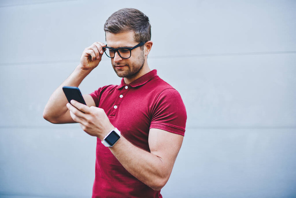 Thoughtful man with beard in trendy outfit and wristwatch touching eyeglasses and browsing smartphone against gray wall of building in daylight - Photo, Image
