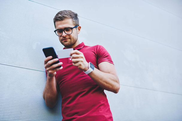 From below of concentrated bearded man in casual clothes and eyeglasses leaning on gray wall and dialing phone number from calling card on smartphone - Fotoğraf, Görsel
