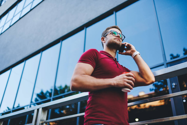 Low angle of pensive man in casual clothes and eyeglasses standing on street and talking on smartphone against contemporary glass building in city in daytime - Zdjęcie, obraz