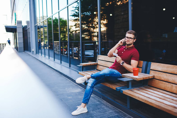 Confident bearded guy wearing casual clothes and eyeglasses sitting on wooden bench with takeaway coffee and reading phone number from business card while making call - Valokuva, kuva