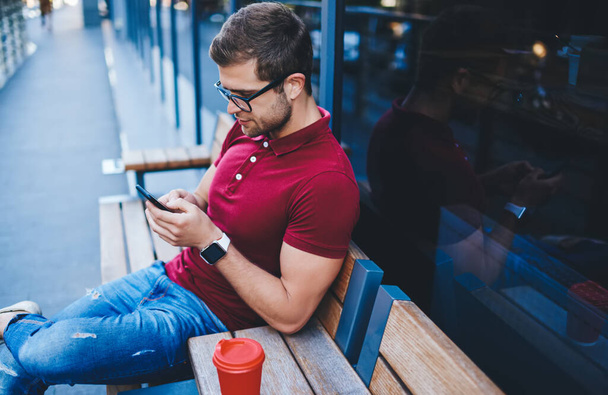 From above of thoughtful bearded guy wearing Polo shirt sitting with red cup of coffee on wooden bench and surfing smartphone on urban background - Foto, imagen
