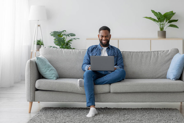 Счастливый молодой черный бородатый мужчина болтает в социальной сети, бизнесмен работает на ноутбуке на диване - Фото, изображение