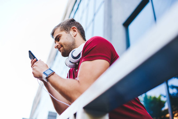 From below of positive young unshaven guy with smart watch and headphones around neck browsing smartphone while leaning on metal railing - Photo, image