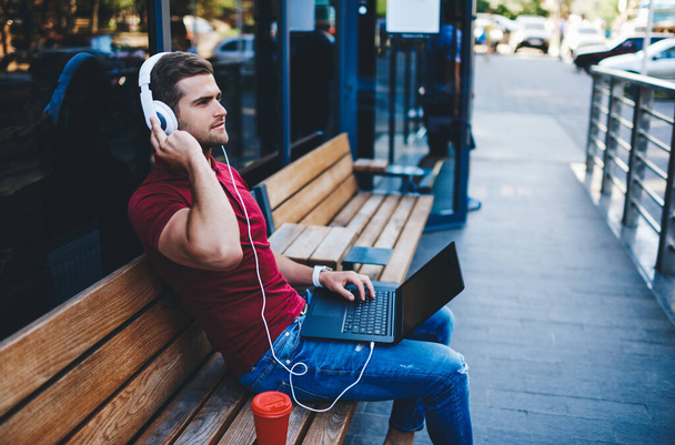 Focused guy in casual outfit sitting on wooden bench near entrance door and listening to audio book in earphones on laptop - Foto, imagen