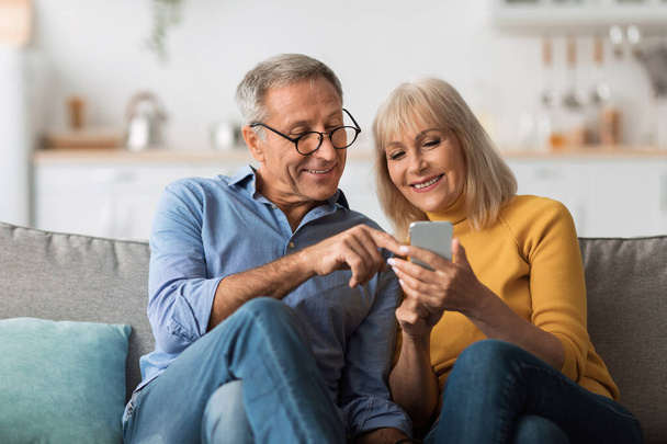 Los cónyuges mayores usan el teléfono móvil sentado en el sofá en casa - Foto, Imagen