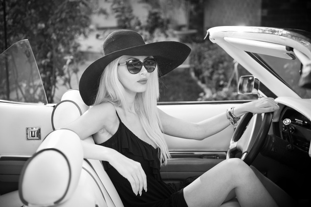 Outdoor summer portrait of stylish blonde vintage woman driving a convertible retro car. Fashionable attractive fair hair female with black hat in withe leather vehicle. Black and white outdoors shot. - Foto, Imagem
