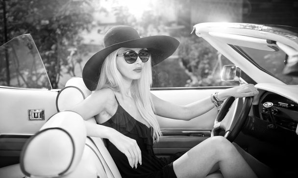 Outdoor summer portrait of stylish blonde vintage woman driving a convertible retro car. Fashionable attractive fair hair female with black hat in withe leather vehicle. Black and white outdoors shot. - Foto, imagen