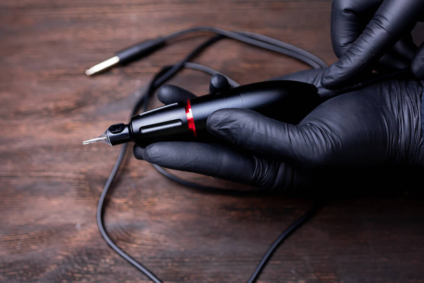 τα χέρια ενός μόνιμου καλλιτέχνη μακιγιάζ με μαύρα αποστειρωμένα γάντια κρατούν ένα μόνιμο μηχάνημα τατουάζ στην παλάμη του χεριού σας - Φωτογραφία, εικόνα