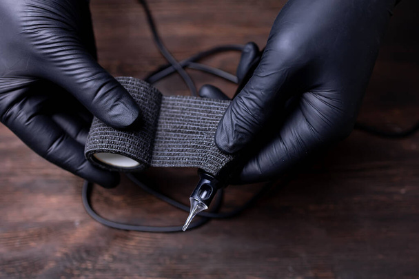 τα χέρια ενός μόνιμου μακιγιέρ με μαύρα αποστειρωμένα γάντια ανεμίζουν μια μαύρη ταινία φραγμού σε ένα μηχάνημα τατουάζ για προστασία - Φωτογραφία, εικόνα