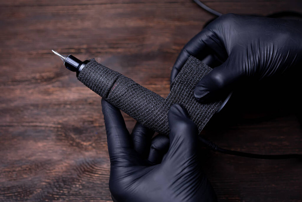 as mãos do mestre de maquiagem permanente em luvas estéreis pretas envolveram uma máquina de tatuagem com uma fita adesiva e cor preta e tentam arrancá-la. - Foto, Imagem