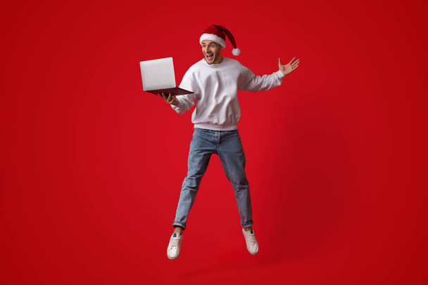Νίκη στο διαδίκτυο. Ευτυχισμένος ενθουσιασμένος άνθρωπος σε Σάντα καπέλο Άλμα με το laptop - Φωτογραφία, εικόνα