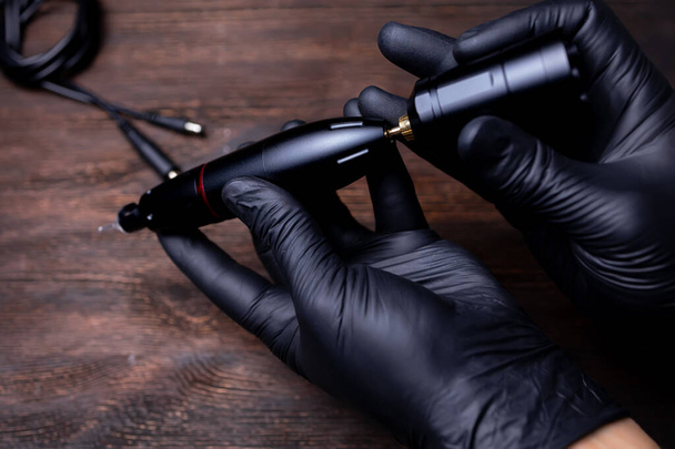 de handen van de permanente make-up kunstenaar in zwarte steriele handschoenen, probeert hij de draadloze batterij pack van de tattoo machine aan te sluiten - Foto, afbeelding