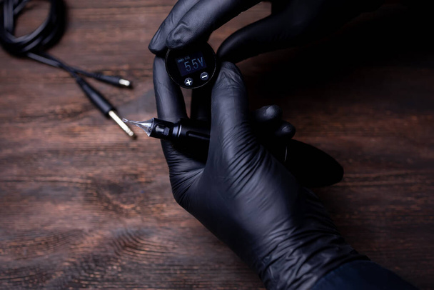 Ręce stałego makijażysty w czarnych jałowych rękawiczkach próbują ustawić napięcie w bezprzewodowym bloku z maszyny do tatuażu. - Zdjęcie, obraz