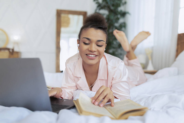 Millennial μαύρη κυρία ξαπλωμένη στο κρεβάτι με φορητό υπολογιστή, διαβάζοντας βιβλίο και έχοντας online τάξη μέσω υπολογιστή, μελετώντας από το σπίτι - Φωτογραφία, εικόνα