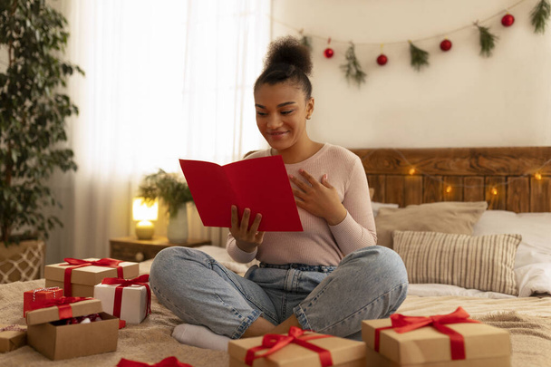 クリスマスおめでとうございます。幸せな黒の女性グリーティングカードを読んでギフトボックスを受け取り、ベッドの上に座って - 写真・画像