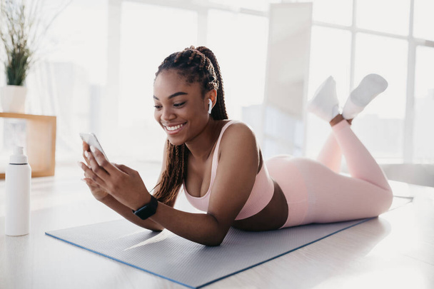 Portrait de jeune femme noire heureuse utilisant un smartphone, écoutant de la musique pendant la pause de la formation domestique - Photo, image