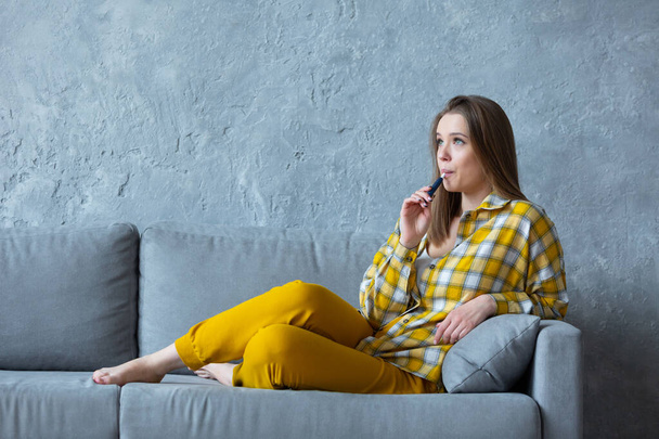 Profile photo of a smiling beautiful girl with an electronic cigarette in her mouth resting on a blue sofa at home - Φωτογραφία, εικόνα