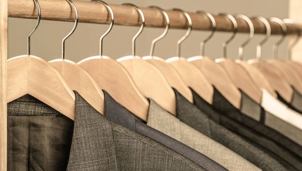 Mens suits in different colors hanging on hanger in a retail clothes store, close-up. Mens shirts, suit hanging on rack. Hangers with jackets on them in boutique. Suits for men hanging on the rack - Photo, Image