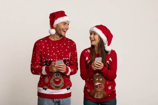 Noel Baba şapkalı neşeli çift ellerinde akıllı telefonlar ve birbirlerine bakıyorlar. - Fotoğraf, Görsel