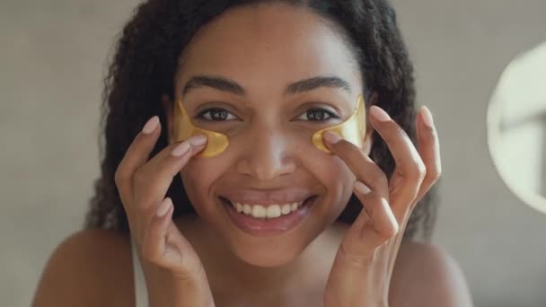 Peili näkökulma muotokuva nuori positiivinen afrikkalainen amerikkalainen nainen laittaa kollageenin laastaria silmien alla alue kylpyhuoneessa - Materiaali, video