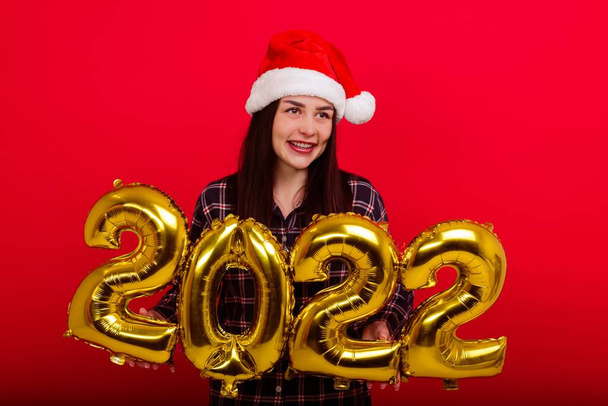 Joulu, loma - nuori hymyilevä nainen pyjamassa ja joulupukin hattu pitävät numeroita 2022 - Valokuva, kuva