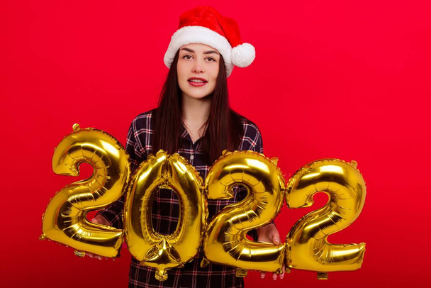 Christmas , concept of a happy holiday - a young woman in pajamas and a Santa Claus hat holds the numbers 2022 - 写真・画像