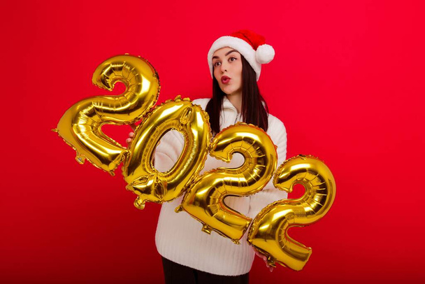 Mutlu tatiller. Kırmızı Noel şapkalı genç bir kadın 2022 rakamlarını taşıyor. - Fotoğraf, Görsel