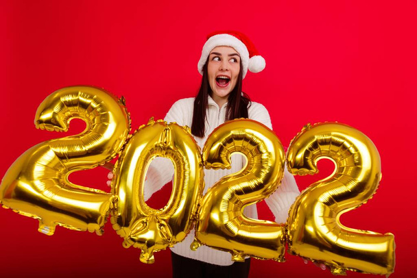 Felices Fiestas - una joven con un sombrero rojo de Navidad sostiene los números 2022 - Foto, Imagen