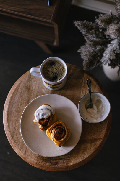 Ψητά ψωμάκια κανέλας με λευκή κρέμα φασολιών βανίλιας. Moody σκοτεινή φωτογραφία τροφίμων. Καφέ και πρωινό με γλυκά. Φρέσκο γλυκό. Καφέ ζάχαρη κανέλας. Στριφογυρίζει σε ρολό. Τραπεζάκι του καφέ. - Φωτογραφία, εικόνα