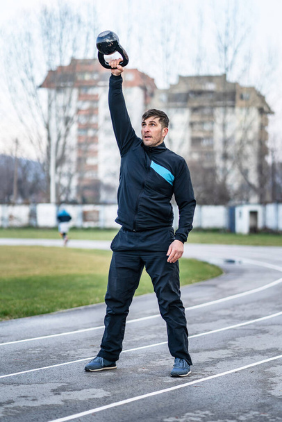 Porträt eines erwachsenen kaukasischen Mannes, der draußen auf der Stadionbahn steht und die Kettlebell in der Hand über dem Kopf hält - Kraft- und Ausdauertrainingskonzept im Frühling oder Herbst - Foto, Bild