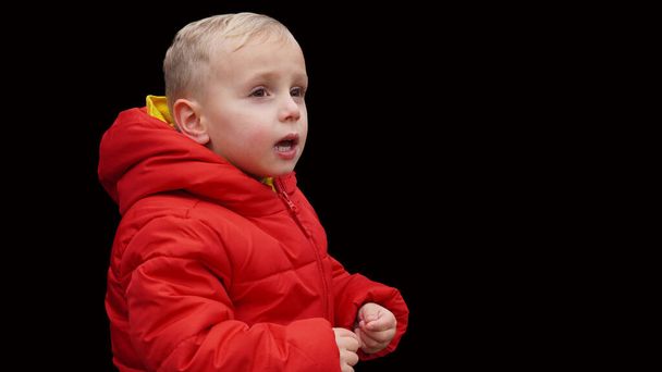 blond kind, in een rode jas op een zwarte achtergrond, drukt zijn emoties - Foto, afbeelding