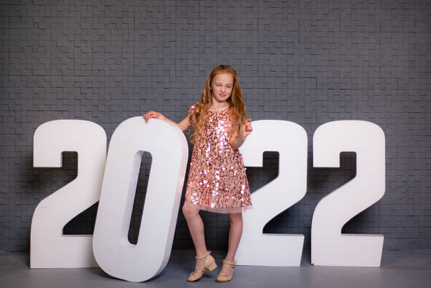 girl and numbers 2022, teenage girl in a shiny dress on a gray brick background with big numbers 2022, New Year's photo with a place for an inscription, copy space - Fotografie, Obrázek