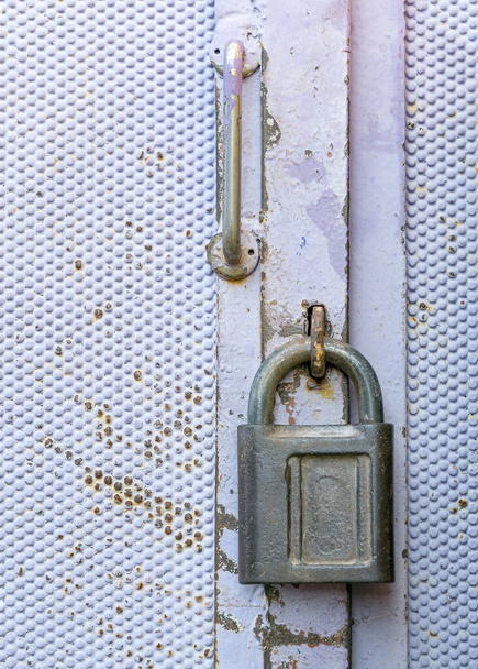 The padlock is hanging on the metal door. The lock shackle is placed in the hinges of the door. The lock is in a closed state. A metal door handle is installed above the lock. - Fotografie, Obrázek