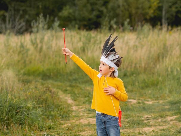 Little boy is playing American Indian on field. Kid has handmade headdress made of feathers and bow with arrows. Costume role play. Outdoor leisure activity. Fall season. - Фото, изображение