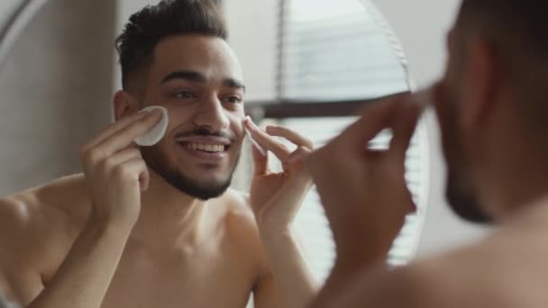 Μεγέθυνση στο πορτρέτο του νεαρού όμορφου καλοφτιαγμένου άντρα που καθαρίζει το πρόσωπό του με ενυδατική λοσιόν και δύο βαμβακερά μαξιλαράκια - Πλάνα, βίντεο