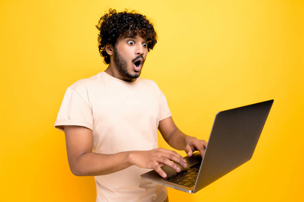 Shocked amazed indian or arabic curly-haired guy, typing on a laptop, looking in surprise at the screen, saw unexpected news, victory, profit, stands on isolated orange background. Emotional face - Photo, image
