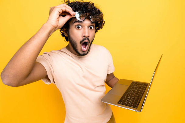 Amazed happy excited Indian or Arabian guy, in casual shirt, take off glasses, holds an open laptop in hand, looks surprised and happily at the camera while standing on isolated orange background - Foto, imagen