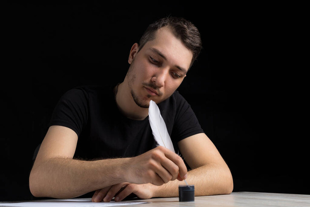 Young shy and smiling handsome man in black clothes dips a quill pen into an inkwell on a black background in a low key - Photo, Image