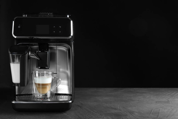 Modern coffee machine with glass cup of latte on grey table against black background. Space for text - Valokuva, kuva