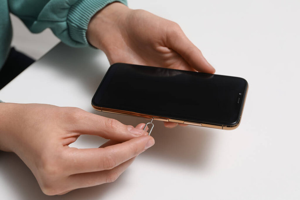 Woman with ejector opening SIM card tray in smartphone at white table, closeup - Foto, Imagem