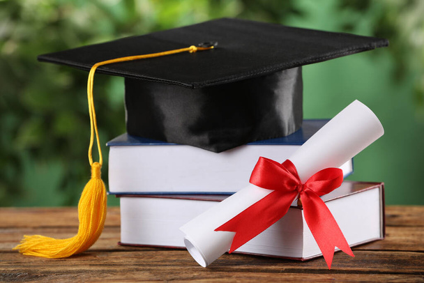 Graduation hat, books and diploma on wooden table against blurred background - Foto, Imagen
