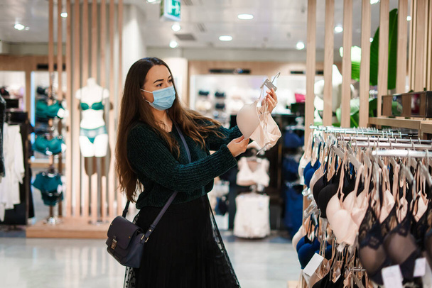 Молода красива жінка в масці для обличчя стоїть у відділі магазину білизни, вибираючи бюстгальтер
 - Фото, зображення