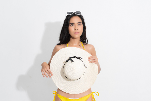 Summer woman studio portrait. Asian woman wearing a yellow bikini dress in a standing position, holding a white white hat and sunglasses, in a summer fashion on isolated white background. - Photo, Image