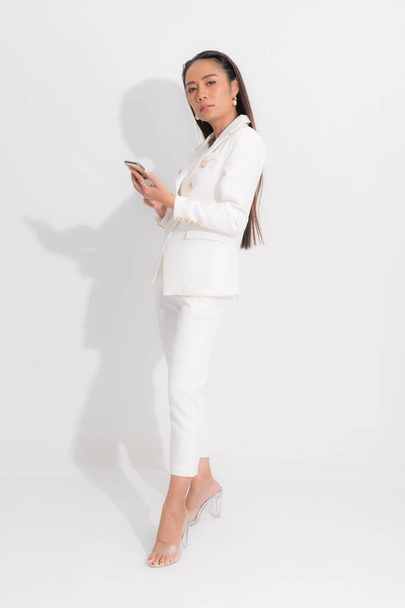 Muoti tyyli luettelo vaatteet liiketoiminnan nainen musta pitkät hiukset luonnollinen muodostavat kulumista valkoinen puku puku täydellinen kehon muoto puku, matkapuhelimella studiossa ampua valkoisella taustalla ja varjossa. - Valokuva, kuva