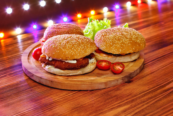 Sortiment tří různých hamburgerů, připravených ke hře, s tmavým dřevěným pozadím a barevnými světly. - Fotografie, Obrázek
